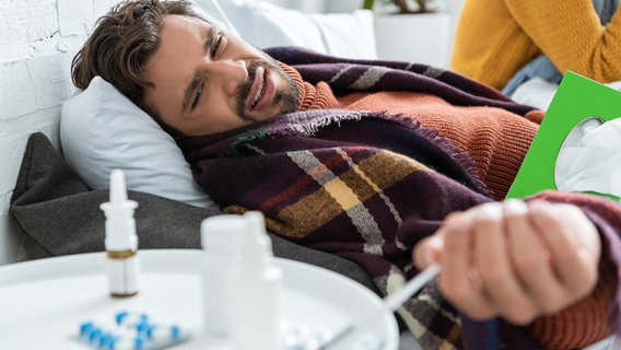 Mann mit schmerzverzerrtem Gesicht liegt mit Schal im Bett vor Nachttisch voller Erkältungsmittel (Bild: colourbox.de) © colourbox.de 