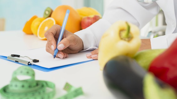 Stoffwechseldiät, Symbolbild: Person mit Arztkittel schreibt auf eine Kladde, daneben liegen ein Maßband, Obst und Gemüse. © IMAGO/ imageBROKER Foto: Oleksandr Latk