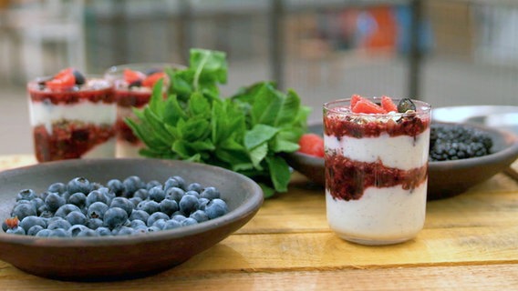 Vanillequark, Beerenmus und Müsli in einem Glas angerichtet. Daneben stehen Schalen mit Beeren. © NDR Foto: NDR Screenshot