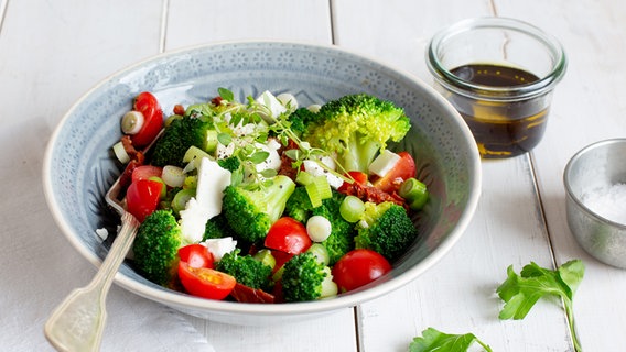 Mediterraner Brokkoli-Salat auf einemTeller. Daneben steht ein kleines Glas Öl-Balsamico-Dressing. © NDR Foto: Claudia Timmann