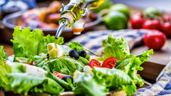 Ein Strahl Olivenöl ergießt sich über eine Schale mit Salat, Tomaten und Oliven © Colourbox Foto: Marian Vejcik