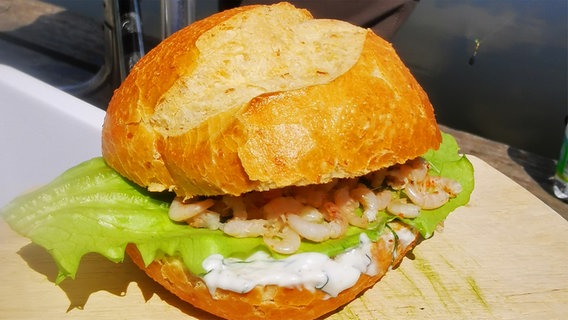 Ein weißes Brötchen mit Krabben, grünem Salatblatt und heller Soße auf einem Holzbrett © NDR Foto: Heinz Galling