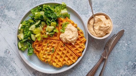 Ein Teller mit einer Waffel und einem grünen Salat, daneben ein Schälchen Hummus. © NDR Foto: Claudia Timmann