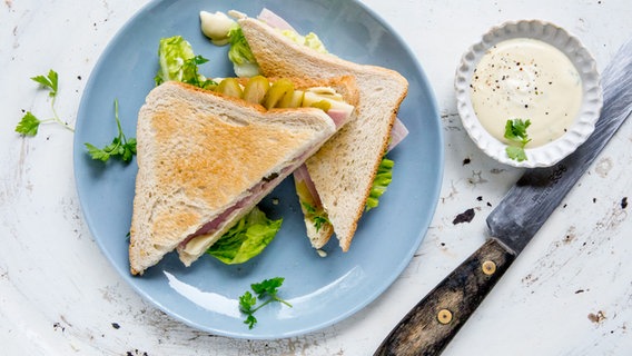 Zwei Käse-Schinken-Sandwiches auf einem Teller mit Honig-Senf-Dip. Daneben liegt ein Holzmesser. © NDR Foto: Claudia Timmann