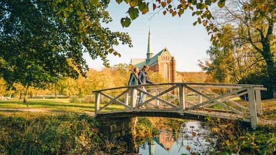 Zwei Spaziergänger gehen über eine kleine Brücke, im Hintergrund das Bad Doberaner Münster. © TMV Foto: Dan Petermann