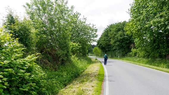 Eine Frau mit Rucksack geht auf einer Straße, die rechts und links von Bäumen und Büschen bewachsen ist. © NDR Foto: Anja Deuble