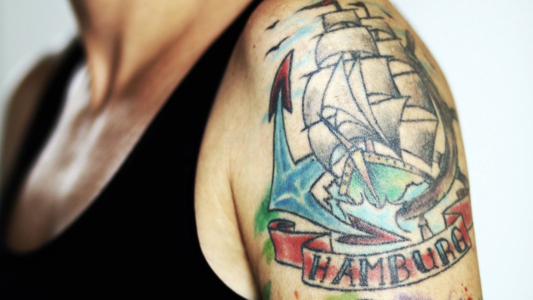 Farbige Tätowierung auf einem Oberarm mit einem Segelschiff und dem Schriftzug Hamburg.