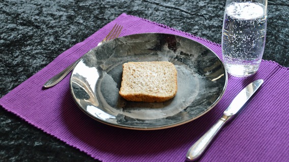 Ein dunkler Teller mit einer Scheibe Brot, daneben Messer und Gabel und ein Glas Wasser © colourbox Foto: Sabine Mederer