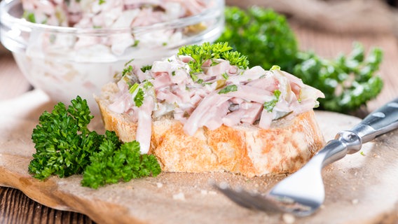 Ein Stück Baguette mit Fleischsalat liegt auch einem Brett aus Holz, daneben eine kleine Schüssel mit Fleischsalat und Petersilie. © picture alliance / Zoonar | Christoph Schöne Foto: Christoph Schöne