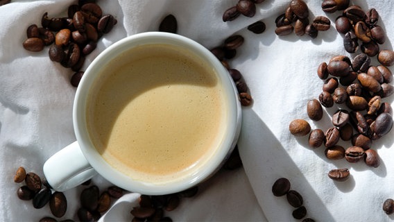 Gefüllte Kaffeetasse von oben auf einer weißen Tischdecke, drum herum liegen einzelne Bohne verstreut. © picture alliance / CHROMORANGE | Alfred Hofer Foto: Alfred Hofer