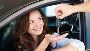 Frau sitzt im Auto und streckt eine Hand aus dem Autofenster, in die ein Autoschlüssel gelegt wird. © Fotolia.com Foto: BillionPhotos.com