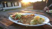 Panierte Scholle liegt mit Kartoffeln und Salat auf einem Teller, im Hintergrund geht die Sonne unter. © picture alliance / augenklick/firo Sportphoto | firo Sportphoto 