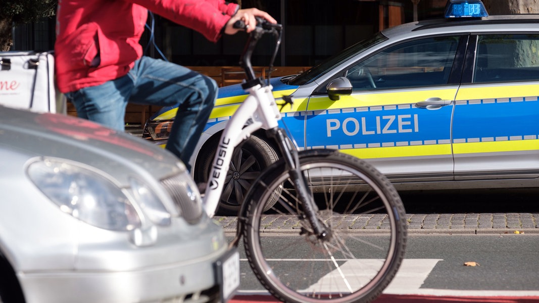 Ein Polizeiauto steht an einer Straße, davor sind ein Auto und ein Radfahrer auf einem Fahrradweg zu sehen.