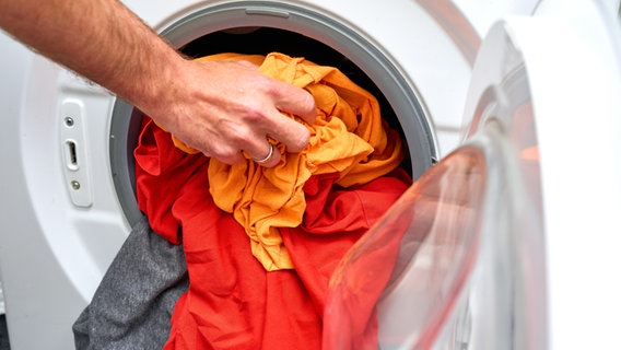 In die Trommel einer Waschmaschine steckt eine Männerhand mehrere T-Shirts in unterschiedlichen Farben. © picture alliance / CHROMORANGE | Michael Bihlmayer Foto: Michael Bihlmayer