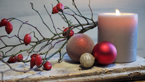 Drei Weihnachtskugeln und ein Zweig mit Hagebutten liegen auf einem alten Brett, daneben steht eine brennende graue Kerze. © NDR Foto: Anja Deuble