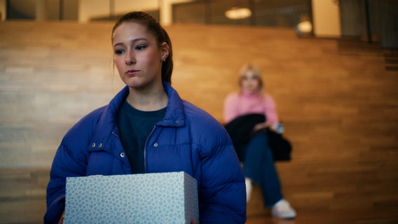 Eine Frau in einer blauen Jacke hält eine gemusterte Schachtel, während sie auf Holzstufen sitzt. Im Hintergrund sitzt eine weitere Person in einem rosa Oberteil, die leicht verschwommen ist. © Mothership 2024/NDR 