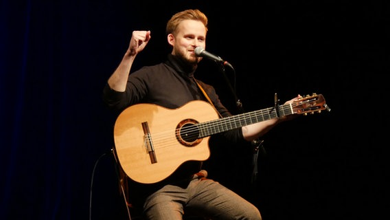 Der Gitarrist Sönke Meinen sitzt mit seiner Gitarre auf einer Bühne und schaut ins Publikum. © NDR Foto: Helgrd Füchsel