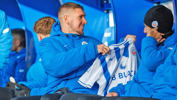 Damian Roßbach vom FC Hansa Rostock hält auf der Ersatzbank ein Trikot in die Höhe © IMAGO / Andy Bünning 