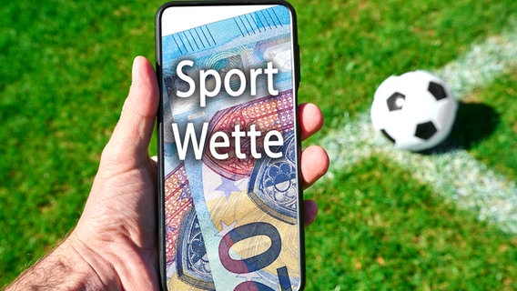 Smartphone mit Aufschrift Sportwette, im Hintergrund liegt ein Fußball © IMAGO / Bihlmayerfotografie 