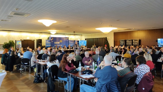 Im Saal des Hotel des Nordens in Harrislee sitzen SSW-Delegierte an Tischen, während Stefan Seidler eine Rede hält. © NDR Foto: Peer-Axel Kroeske