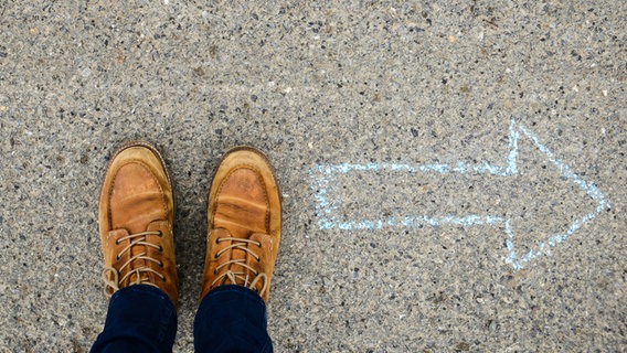 Blick von oben auf Schuhe und Boden, auf dem ein Pfeil, der nach rechts zeigt, mit Kreide auf den Asphalt gezeichnet wurde. © David-W-/photocase Foto: David-W-