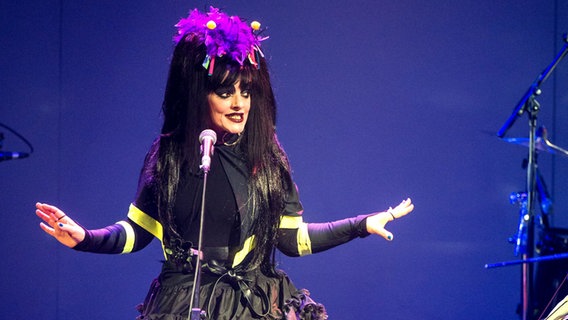 Nina Hagen steht auf einer Bühne hinter einem Mikrofon. Sie trägt ihre schwarzen Haare zu zwei Zöpfen gebunden mit lila Blumen auf dem Kopf und ein schwarzes Outfit mit gelben Bändern dran. © Picture-Alliance / dpa Foto: Bernd von Jutrczenka