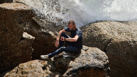 Der Musiker Pohlmann sitzt auf einem Felsen im Wasser und schaut auf die Uhr. © Pohlmann Foto: Benedikt Schnermann