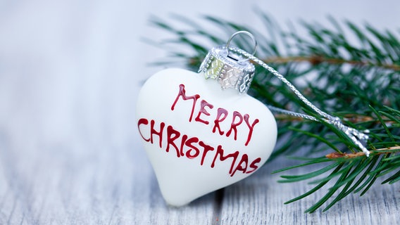 Ein weißes Christbaumherz mit der Aufschrift "Merry Christmas" und ein Tannenzweig liegen auf einem weißen Holztisch. © Colourbox Foto: JuNiArt