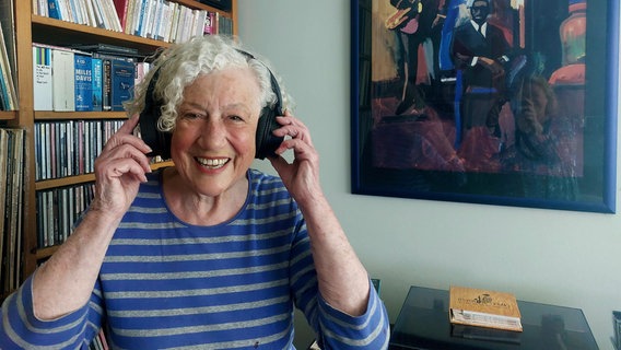 Die Künstler-Promotorin Elfie Küster hat Kopfhörer auf den Ohren und lächelt in die Kamera. © NDR Foto: Mechthild Maesker
