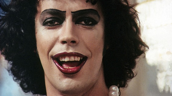 Eine Filmszene aus der Rocky Horro Picture Show: Tim Curry lächelt die Kamera lasziv an. © picture-alliance / KPA 