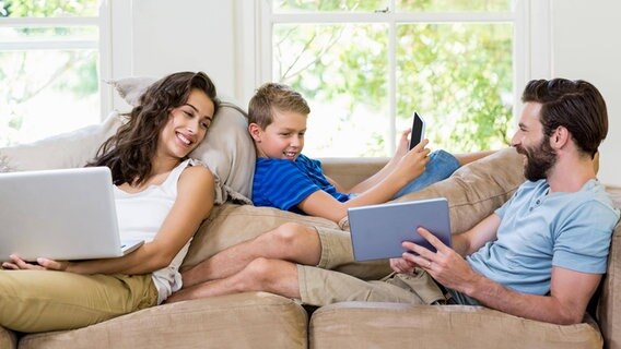 Eltern sitzen zusammen mit ihrem Sohn auf einer Couch und schauen lächeld auf ein Tablet. © WavebreakmediaMicro 