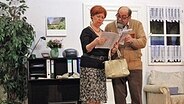 Eine Frau und ein älterer Herr stehen in einer Wohnzimmerkulisse und schauen gemeinsam in die Zeitung © Ursula Visser Foto: Ursula Visser
