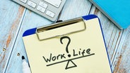 Auf einem Tisch liegt ein Klemmbrett mit einem Blatt Papier auf dem "Work?Life" steht. © Colourbox 